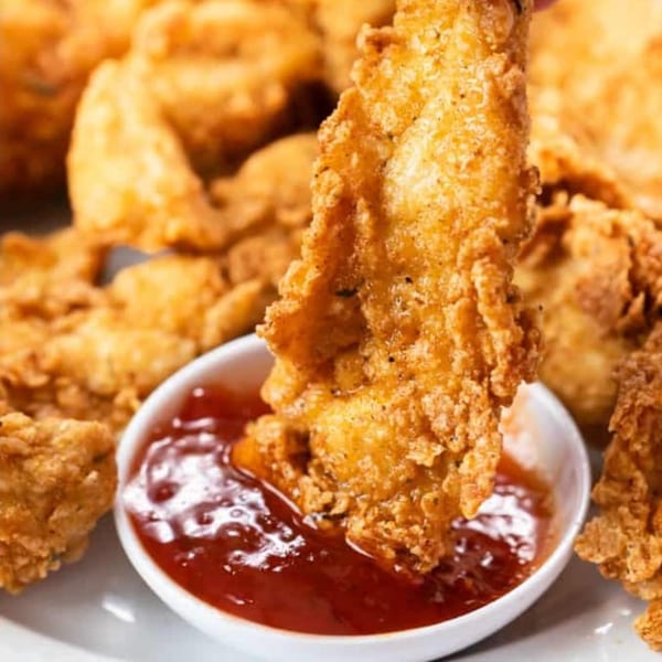 Crispy golden-brown chicken tender being dipped into a small white bowl of red sauce.