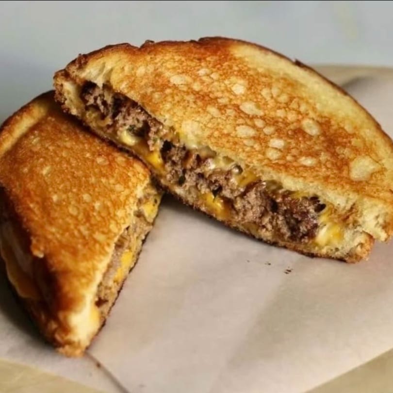 A grilled cheese sandwich with ground beef and melted cheese, cut in half on parchment paper.