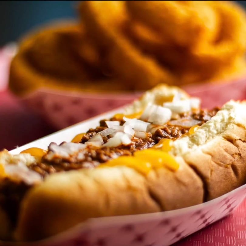 A chili cheese dog with diced onions in a paper tray, with onion rings blurred in the background.