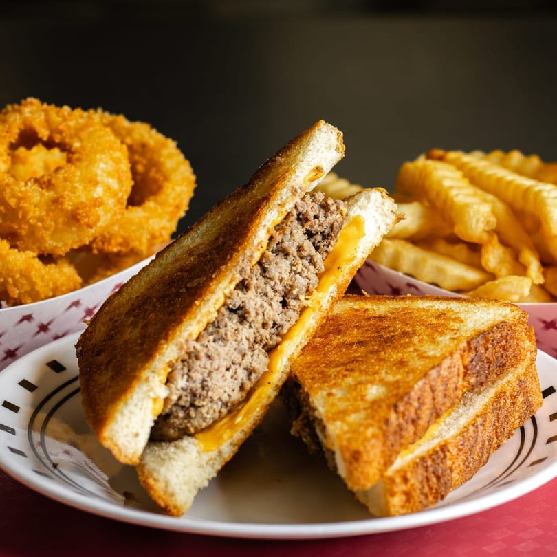 A grilled cheese sandwich with ground beef and melted cheese, served with onion rings and fries.